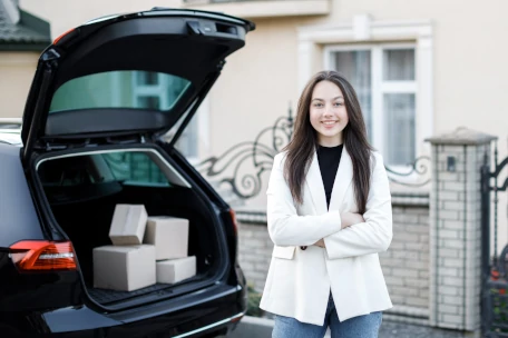 Jeune femme souriante debout devant le coffre ouvert de sa voiture contenant des colis.